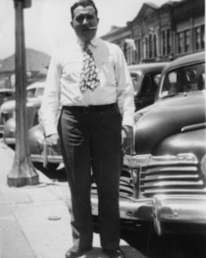 Clyde Smaldone, cigar in mouth, standing on a Denver sidewalk in front of cars sometime in 1940s.